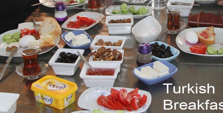 Turkish breakfast dishes arranged on a table with bread, cheese, yogurt, tomatoes, cucumbers, eggs, olives, honey, jam.