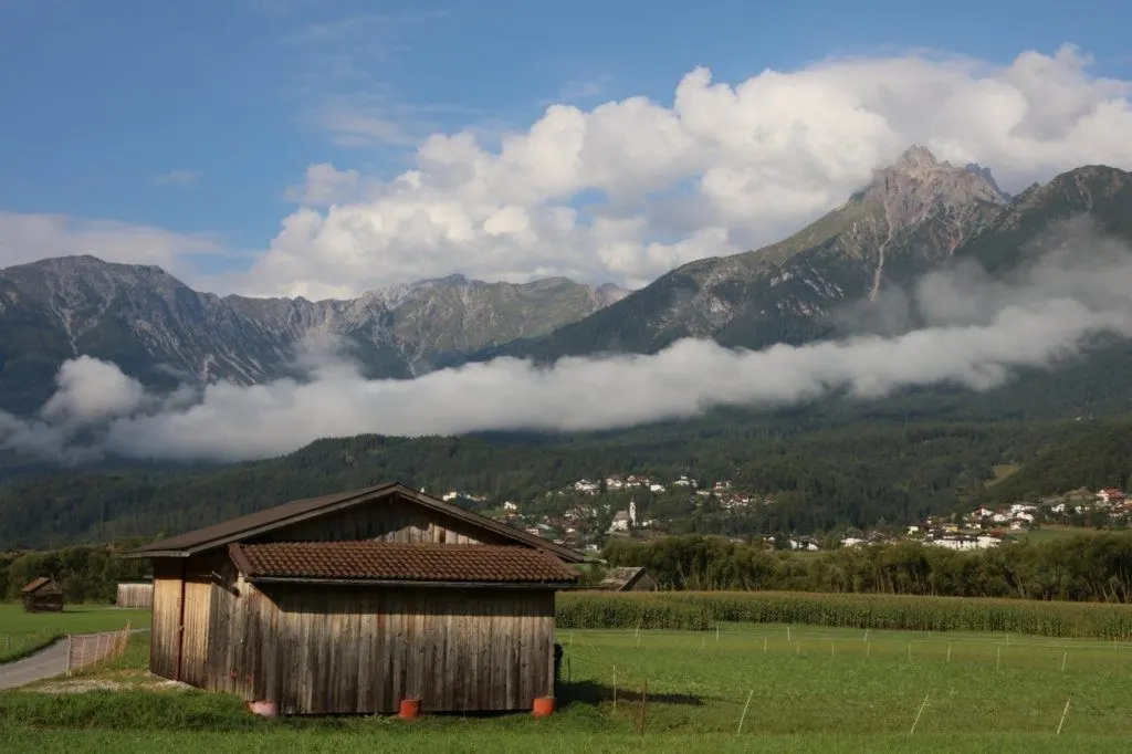 A cow shed in an alpine field with mountain peaks in the distance provides shelter for shepherds.