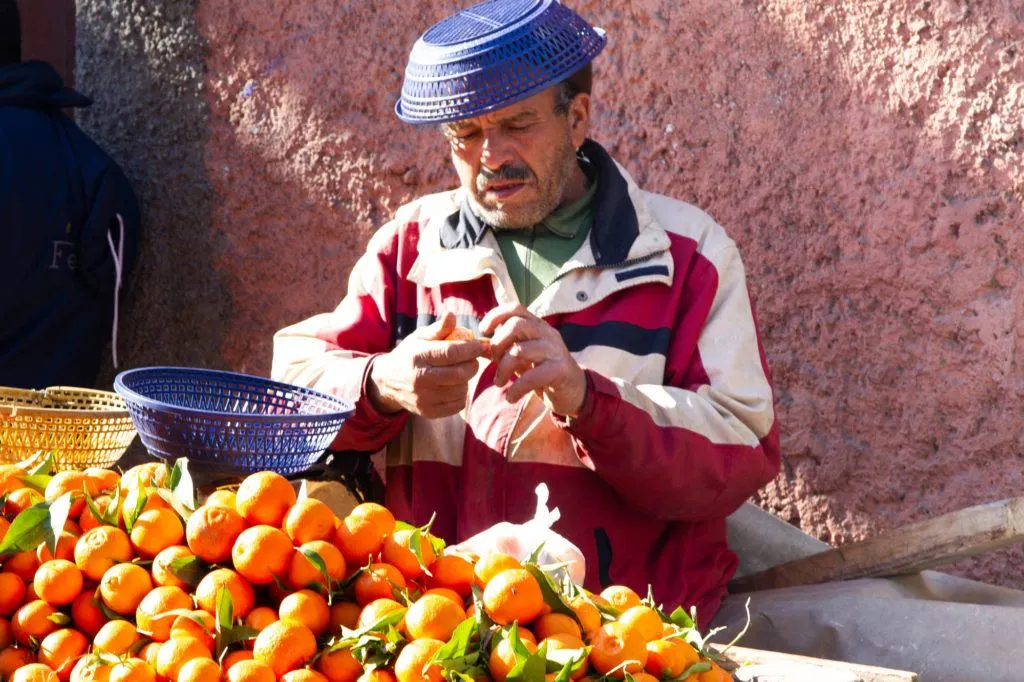 An orange vendor, wearing a blue plastic basket for a hat, sells fresh oranges from a cart in Jemaa el Fna.