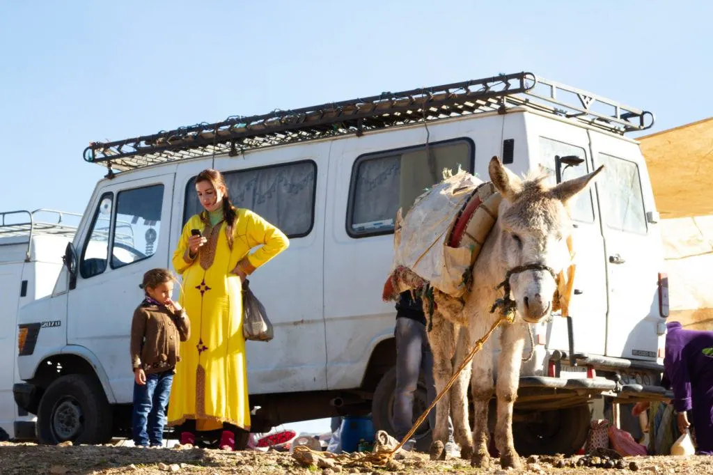 A young woman wearing a bright yellow djellaba is standing near a donkey and looking at her cell phone.