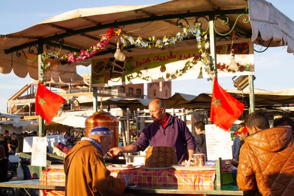 A vendor with his cart decorated with garland and Moroccan flags serves mint tea to a customer.