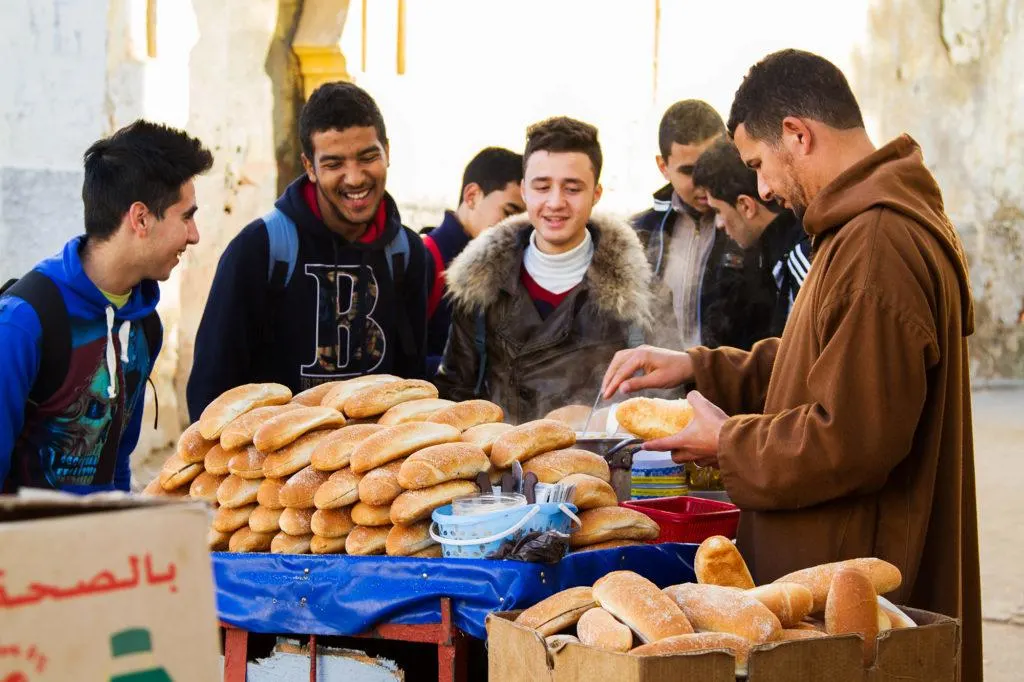 A group of teenage boys at a table stacked with small loves of bread waiting to be served by the vendor.