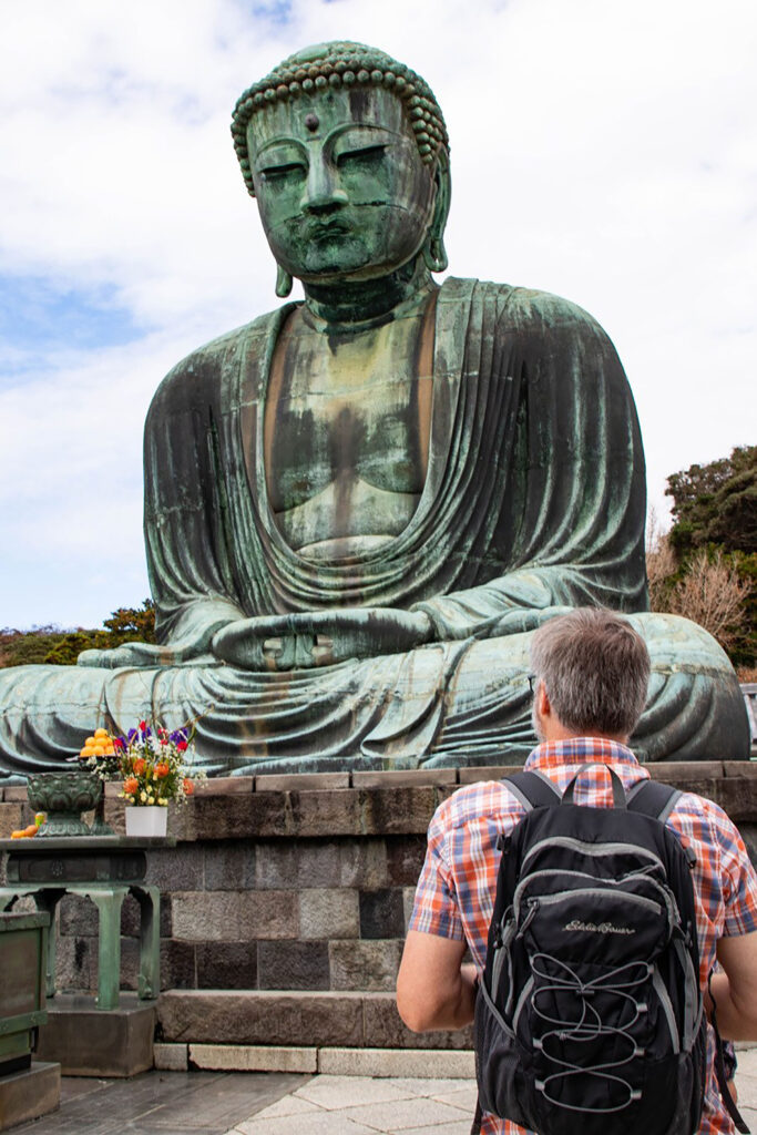A day tripper wearing a day trip bag is standing before the Great Buddha in Kamakura.